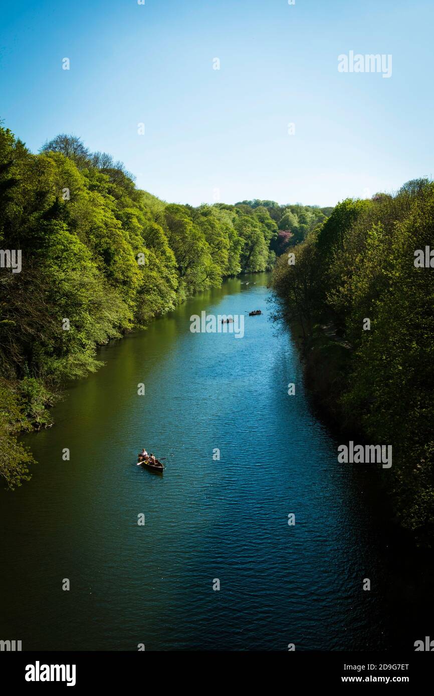 A few boats along a pleasant river. Flanked by thick green trees and basked in a summer suns hazing glare. A single boat towards the foreground pulls. Stock Photo