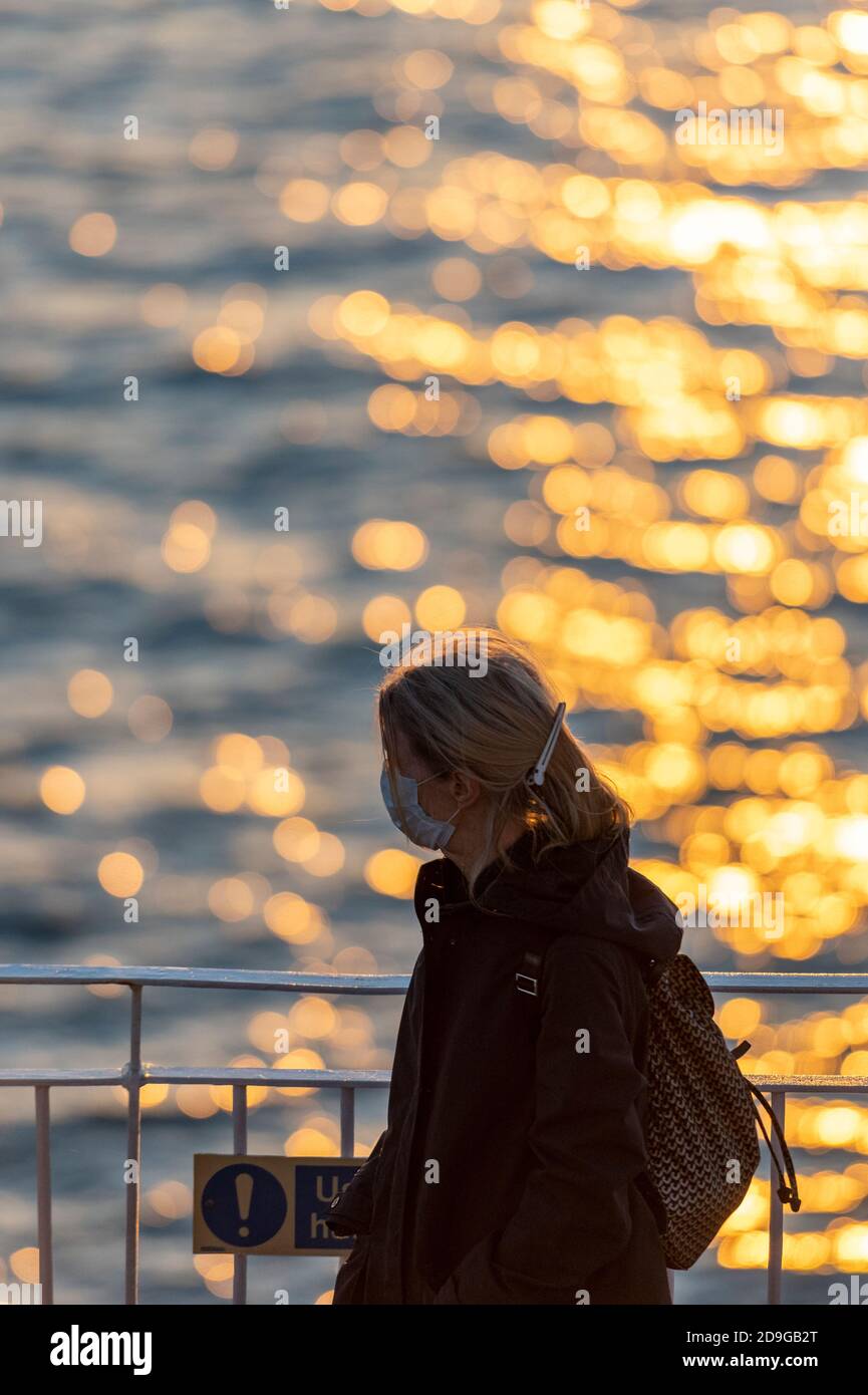 a woman sitting next to some railings next to the sea at sunset looking out over the golden coloured water daydreaming. Stock Photo