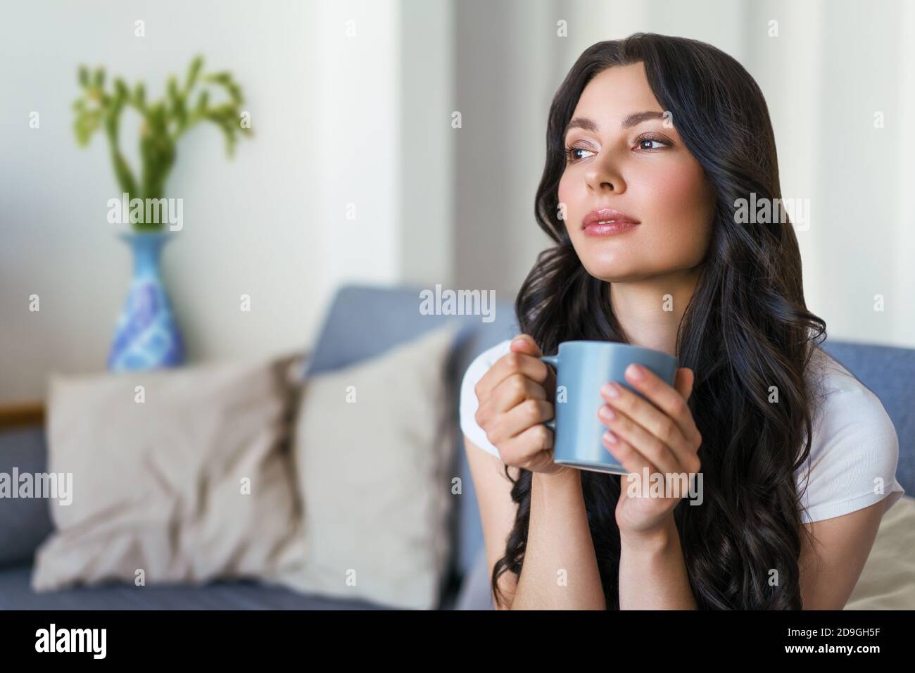 Young happy woman drinking tea and dreaming sitting on the couch in the bright apartment. Stock Photo