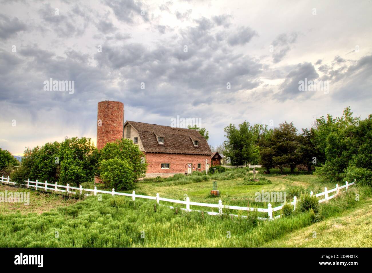 On a cloudy summer day a red brick barn and silo sit in a lush meadow surrounded by a white rail fence. Stock Photo