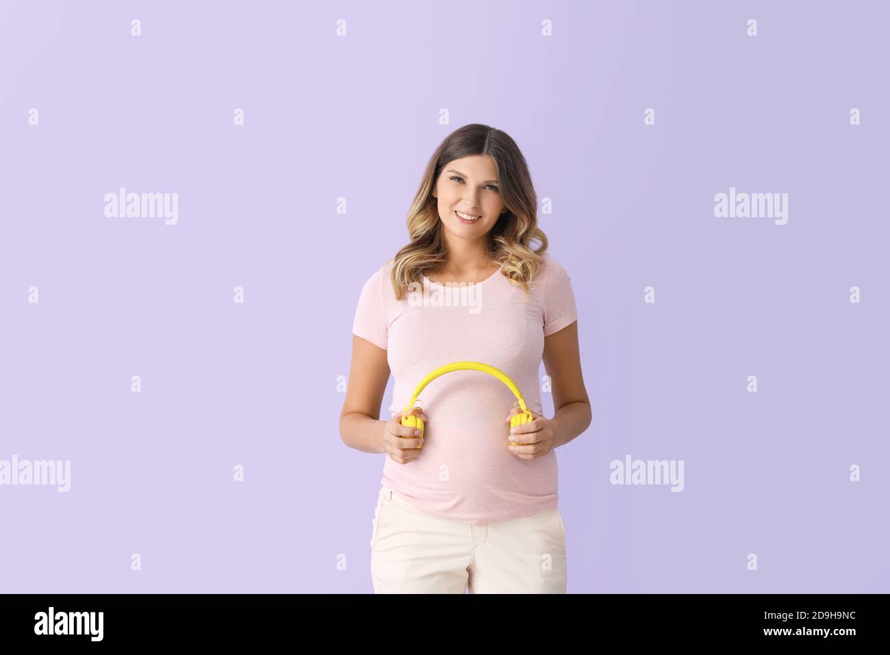 Beautiful pregnant woman putting headphones on her belly against color