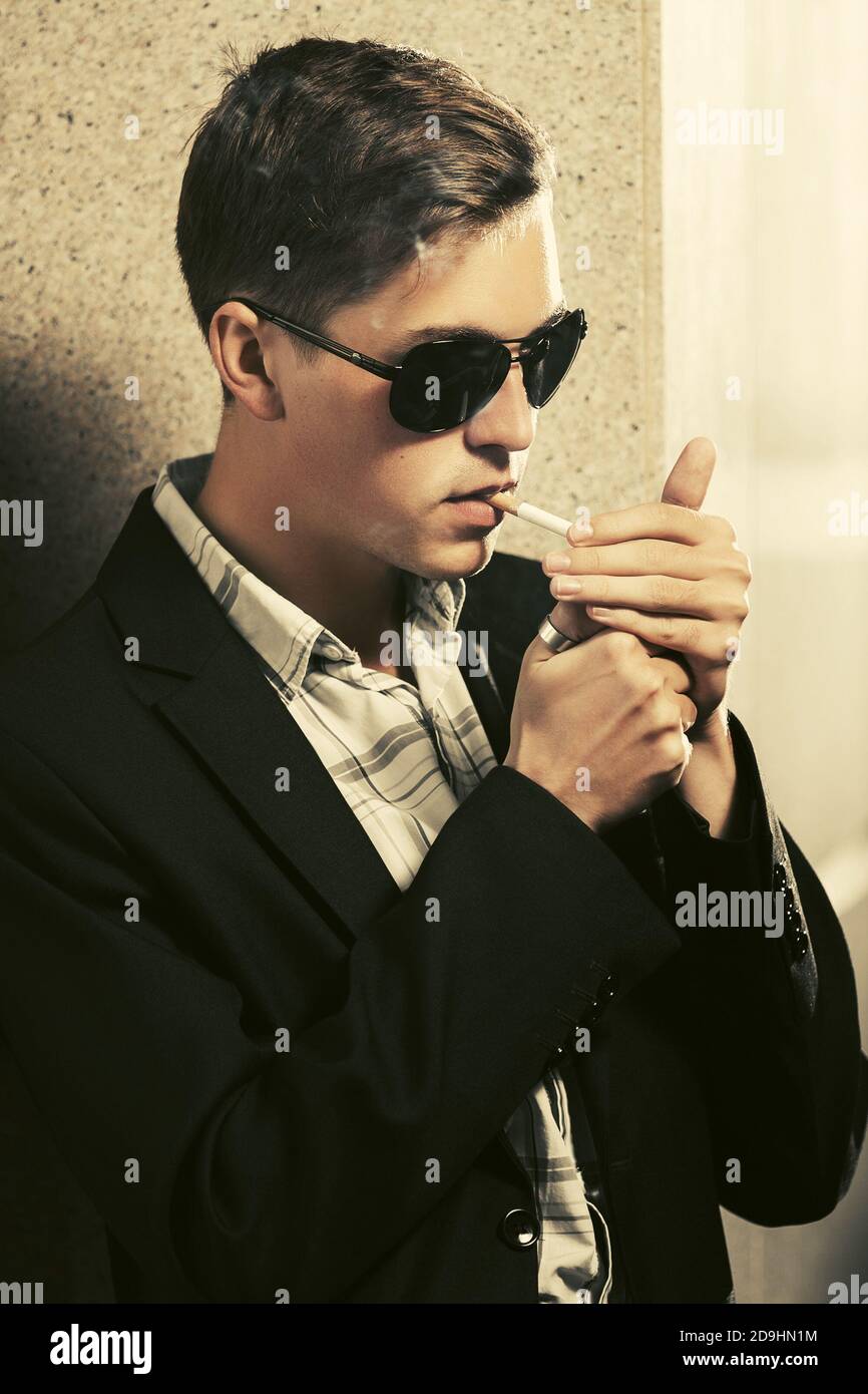 Young man in sunglasses smoking a cigarette on city street  Stylish fashion male model wearing black suit jacket Stock Photo