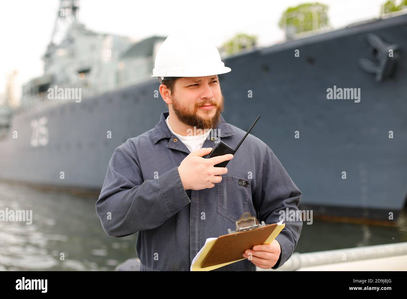 Marine european captain talking by walkie talkie radio and standing near vessel with documents in hands. Stock Photo