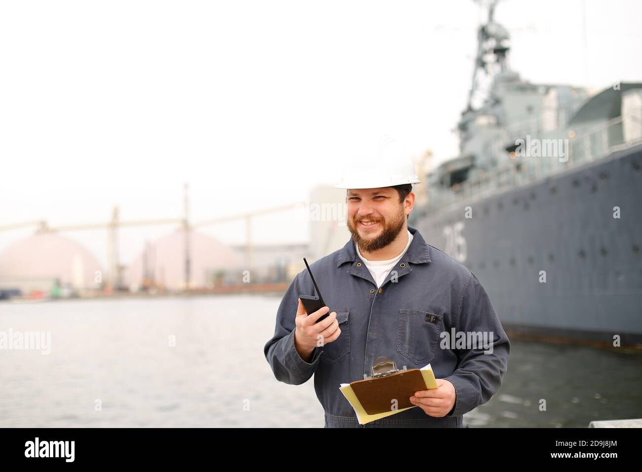 Marine caucasian captain talking by walkie talkie radio and standing near vessel with documents in hands. Stock Photo