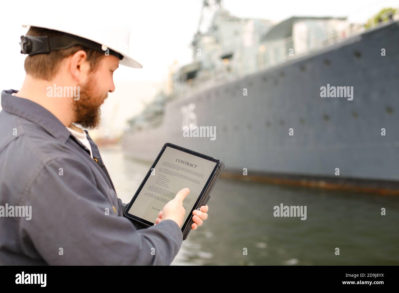 Captain in helmet reading contract on tablet near vessel in background. Stock Photo