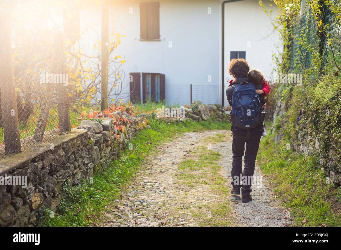 Young father walking and carrying a child girl on a country road scenery with copy space and sunflare. Varese, Italy. Stock Photo
