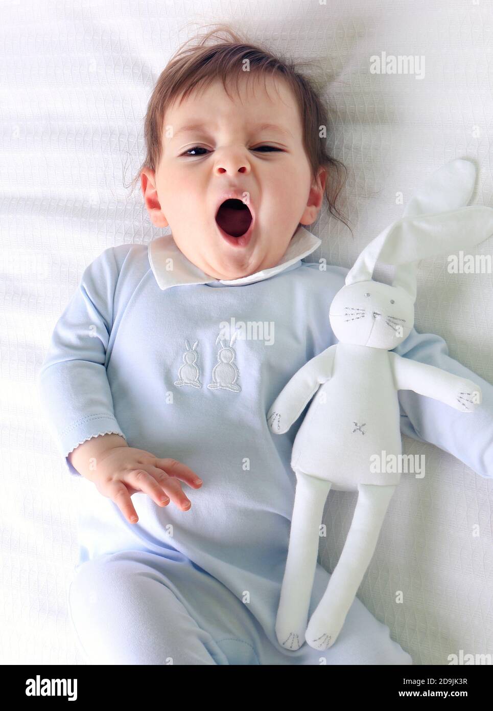 A yawning baby wearing blue and holding his bunny rabbit plush toy. Stock Photo