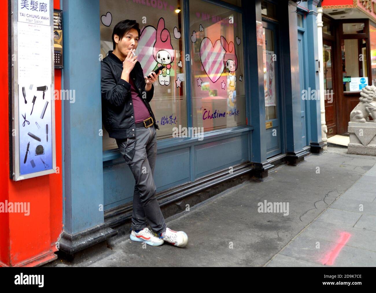London, England, UK. Chinatown: Gerrard Street - young Chinese man looking at his mobile phone and smoking a cigarette Stock Photo