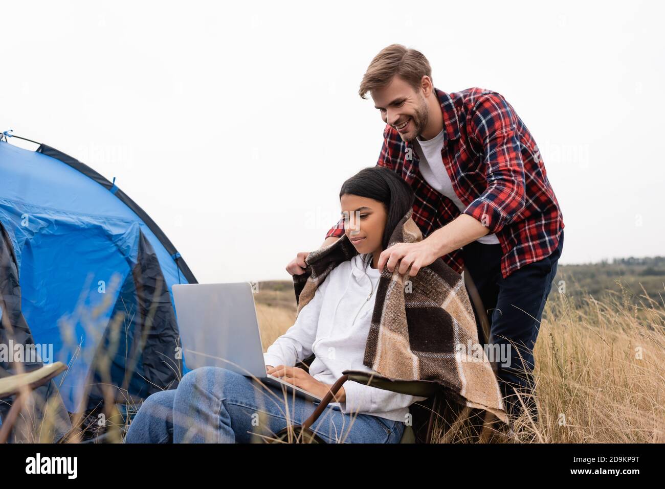 African american woman using laptop near smiling boyfriend with blanket during camping on meadow Stock Photo