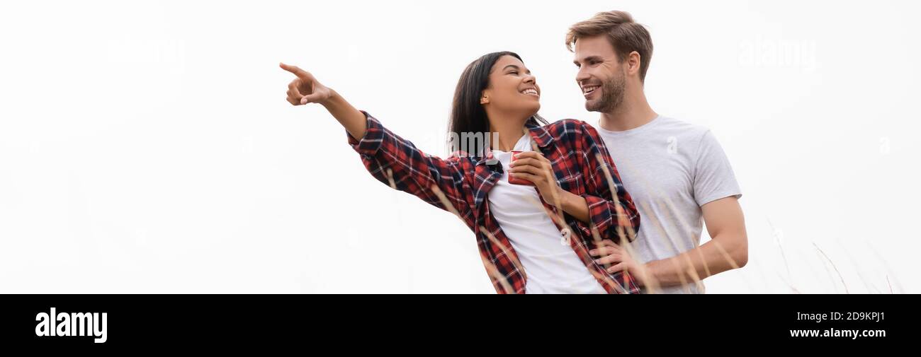 African american woman with cup pointing with finger and smiling at boyfriend near grass on blurred foreground isolated on white, banner Stock Photo
