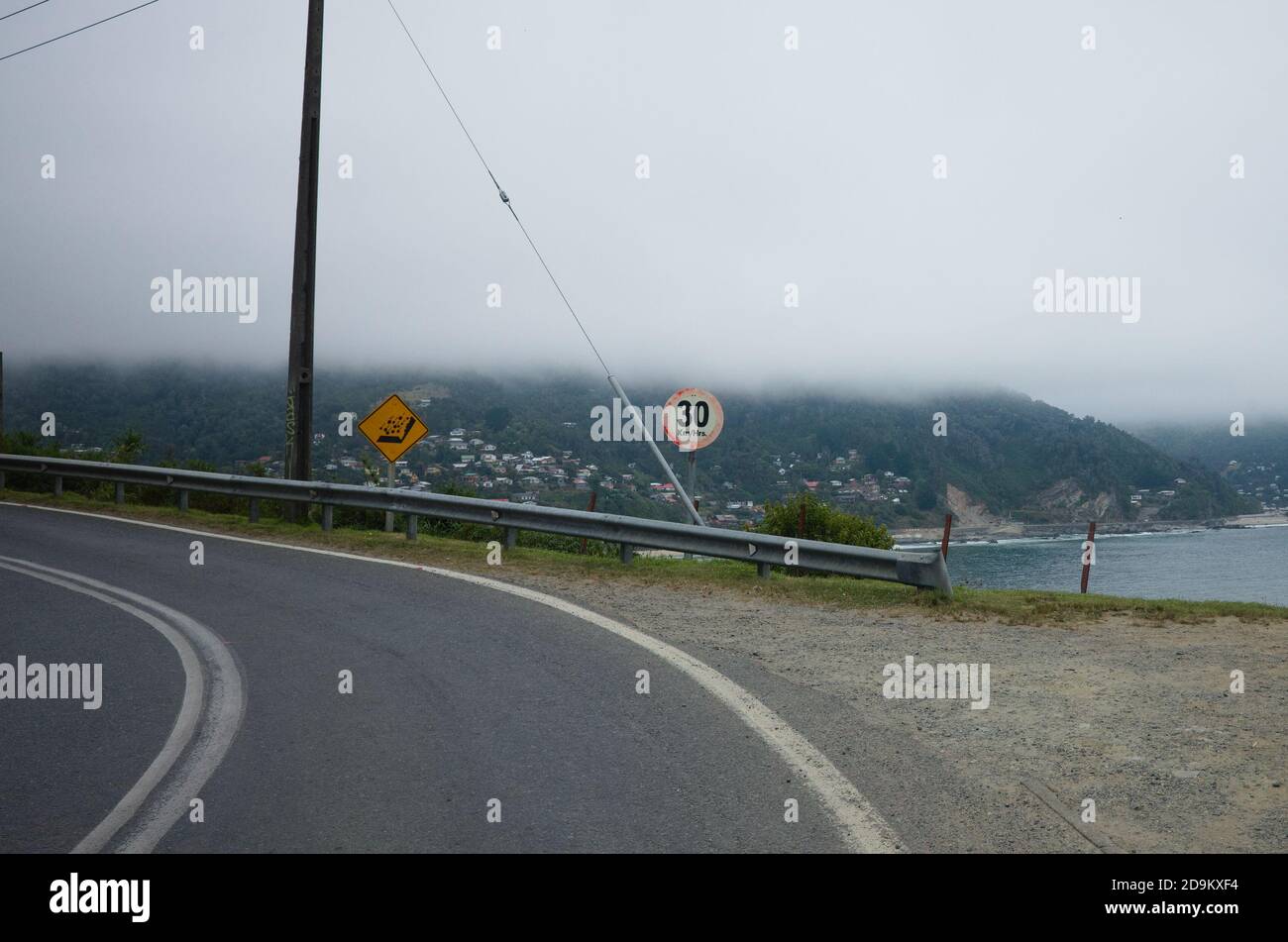 Sharp turn road before cliff and speed limit sign behind. Warning sign falling rocks. Ocean beach and village on background. Chile Stock Photo