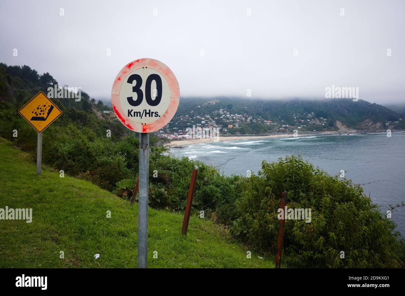 Road sign speed limit on a road in mountains with sharp curve. Rocks falling warning traffic sign. Ocean beach and village on background. Chile Stock Photo