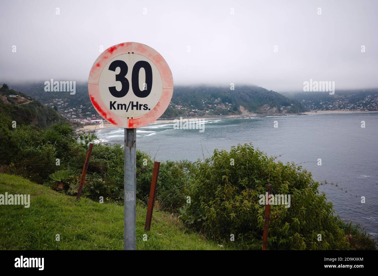 Speed limit sign on a sharp turn before cliff. 30 km per hour speed limit on mountain road. Ocean beach and small village in a fog on background. Stock Photo