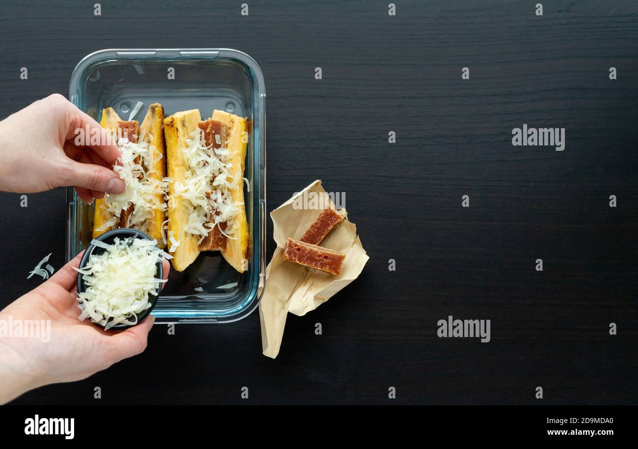 Hand putting cheese to cook Ripe banana in the oven with guava and cheese sandwich on a black wooden base. Typical Latin food concept. Copy space. Stock Photo