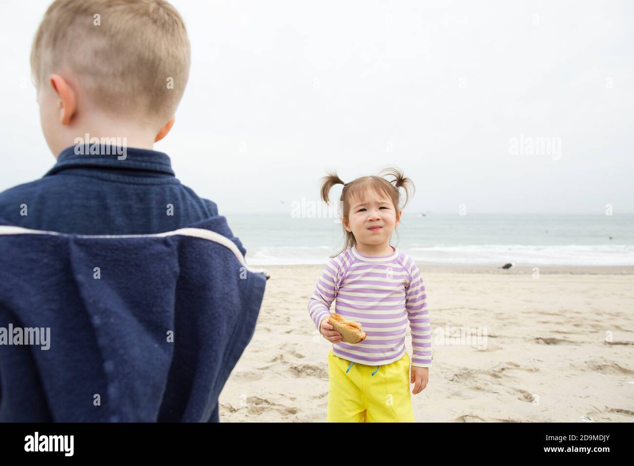 Toddler Girl Stands Eating Peanut Butter Sandwich on Beach Next to Boy