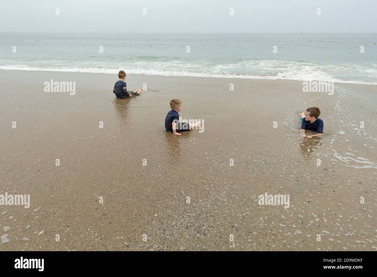 Three Boys Sit and Lie Down at Ocean's Edge While on Beach Vacation Stock Photo
