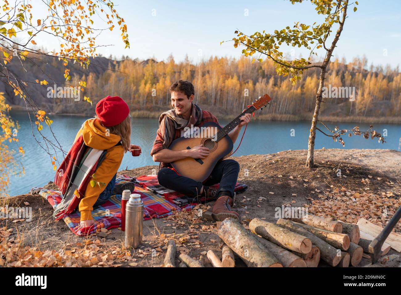 A couple are resting in nature, against the background of autumn trees and the river. Guy plays the guitar Stock Photo