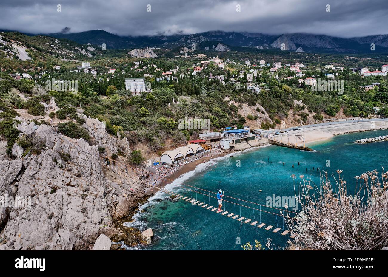 Suspension bridge to the Diva rock, Black Sea coast, near Yalta, Crimea. Male tourist walking on the bridge. Panorama of the town of Simeiz. Stock Photo