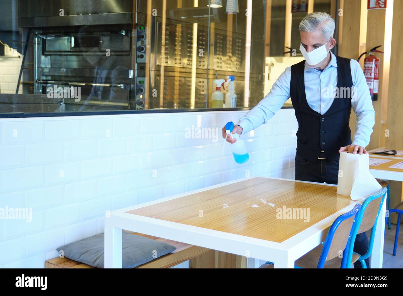 Caucasian male worker cleaning table with disinfectant in a restaurant during coronavirus outbreak. Waiter in protective mask disinfecting table with Stock Photo
