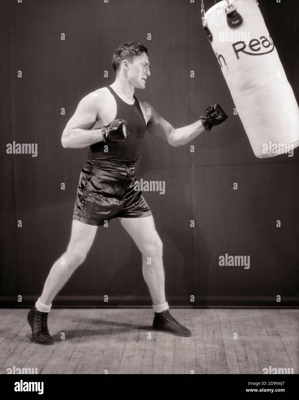 1930s MAN BOXER WEARING BLACK TRUNKS AND TANK SPARRING PRACTICING EXERCISING WITH A REACH HEAVY BAG PUNCHING BAG - g4369 HAR001 HARS COPY SPACE FULL-LENGTH PHYSICAL FITNESS PERSONS INSPIRATION MALES ATHLETIC B&W HITTING GOALS ACTIVITY PHYSICAL STRENGTH AND RECREATION OCCUPATIONS PUNCHING CONNECTION ATHLETES FLEXIBILITY MUSCLES PUGILIST PRIZEFIGHTING FISTICUFFS MID-ADULT MID-ADULT MAN PRECISION SPARRING STRIKING TRUNKS WORKOUT BLACK AND WHITE CAUCASIAN ETHNICITY HAR001 OLD FASHIONED PRACTICING Stock Photo