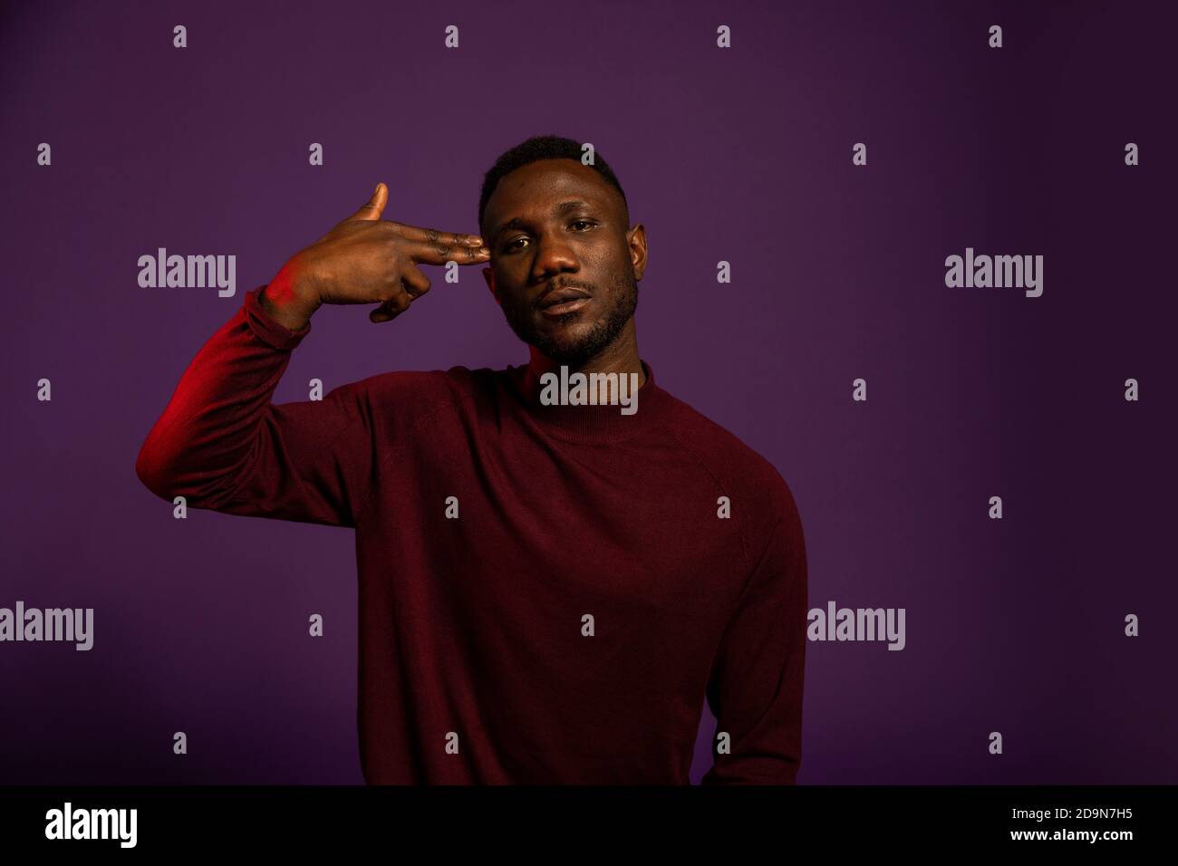 Young black man showing the shoot me hand gesture with fingers to temple. Medium shot. Isolated background. Looking at camera. Stock Photo