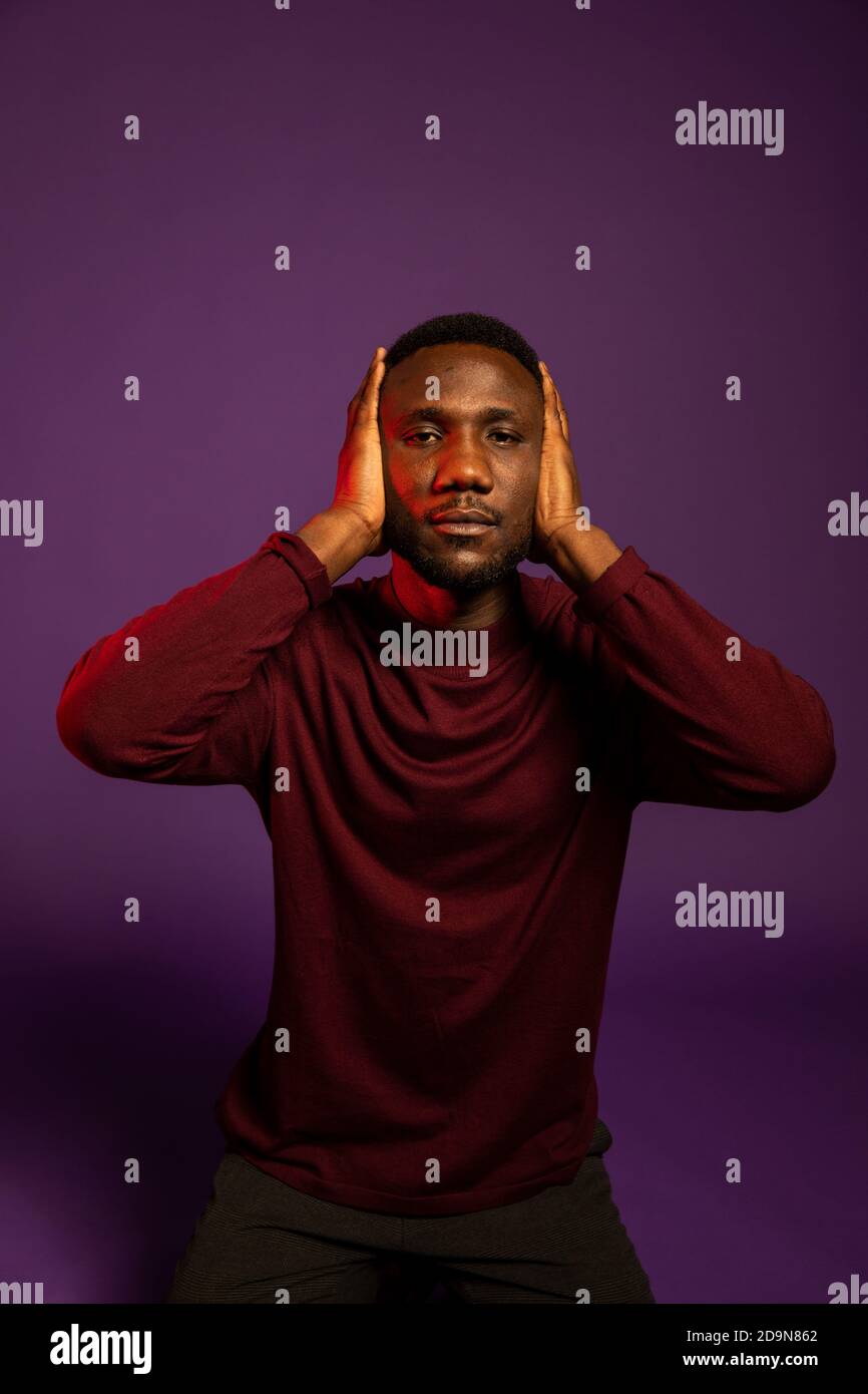 Young black man with hands covering ears looking at camera. Medium shot. Isolated background. Stock Photo