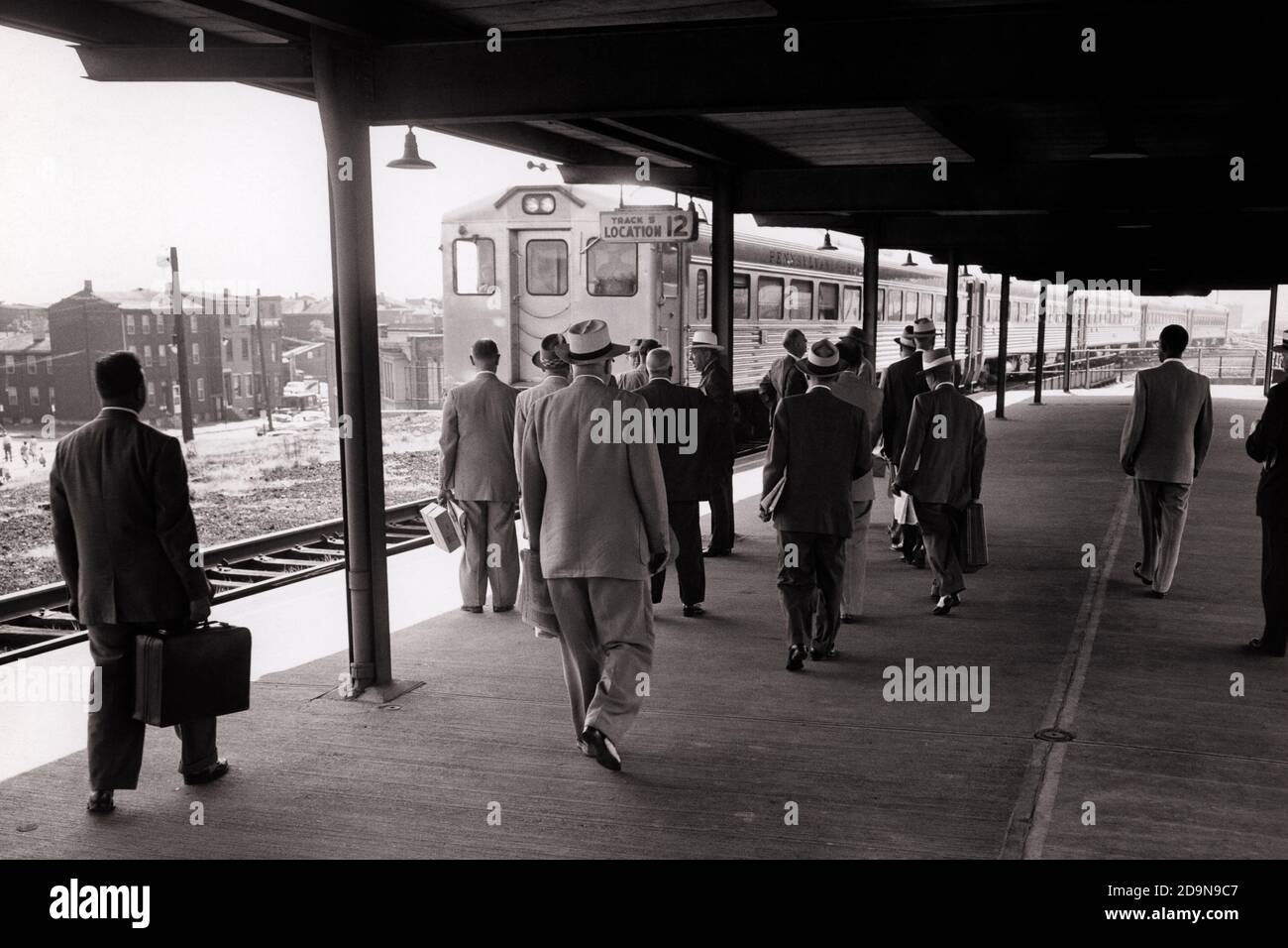 1960s MEN IN SUITS AND HATS BUSINESSMEN BACK VIEW OF SUBURBAN STATION PASSENGERS WAITING FOR REGIONAL COMMUTER TRAIN - r526 HAR001 HARS TRANSPORTATION B&W NORTH AMERICA NORTH AMERICAN RAIL SKILL OCCUPATION PLATFORM SKILLS COMMUTERS AND REAR VIEW IN OF NJ OCCUPATIONS CAMDEN CONNECTION FROM BEHIND NEW JERSEY RAILROADS BACK VIEW COOPERATION TOGETHERNESS BLACK AND WHITE COMMUTER HAR001 OLD FASHIONED REGIONAL Stock Photo