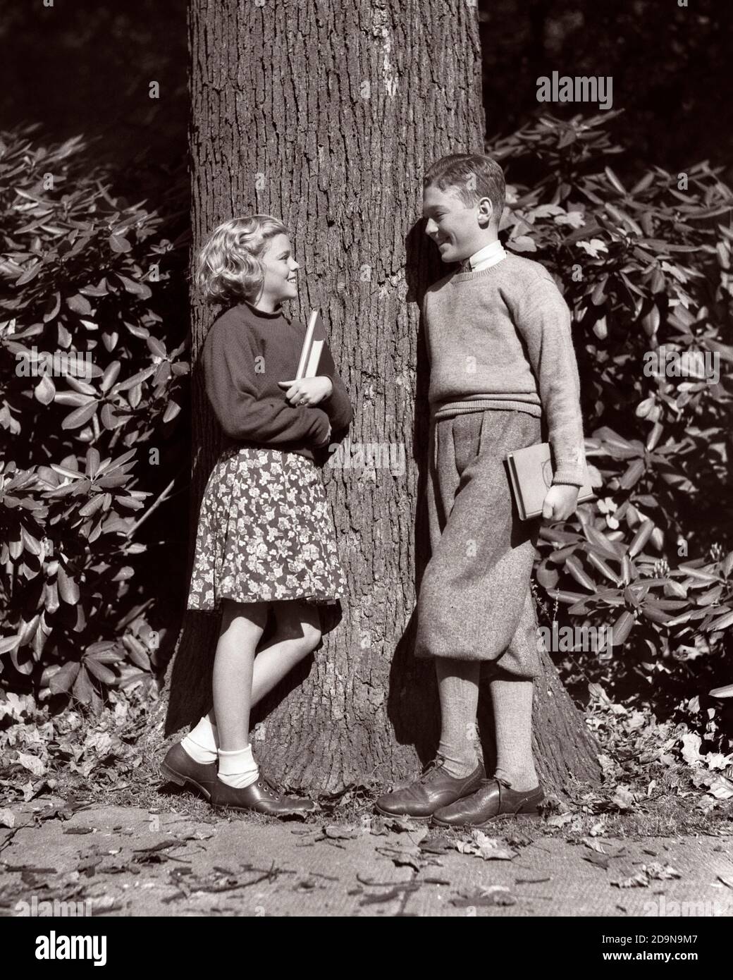 1930s SMILING BOY AND GIRL PRETEENS TEENAGE FRIENDS LEANING AGAINST TREE HOLDING TEXTBOOKS TALKING LISTENING SOCIALLY LEARNING  - s6636 HAR001 HARS 1 JUVENILE STYLE FRIEND JOY LIFESTYLE FEMALES HEALTHINESS COPY SPACE AGAINST FRIENDSHIP FULL-LENGTH PERSONS MALES TEENAGE BOY B&W HAPPINESS AND FEELING PRETEEN TEXTBOOKS FRIENDLY STYLISH TEENAGED CRUSH KNICKERBOCKERS PLUS FOURS PUPPY LOVE AFFECTION CHILDHOOD INFORMAL JUVENILES MIDDLE SCHOOL PLATONIC PRE-TEEN PRE-TEEN GIRL TOGETHERNESS BLACK AND WHITE CAUCASIAN ETHNICITY FIRST LOVE HAR001 IMMATURE OLD FASHIONED Stock Photo