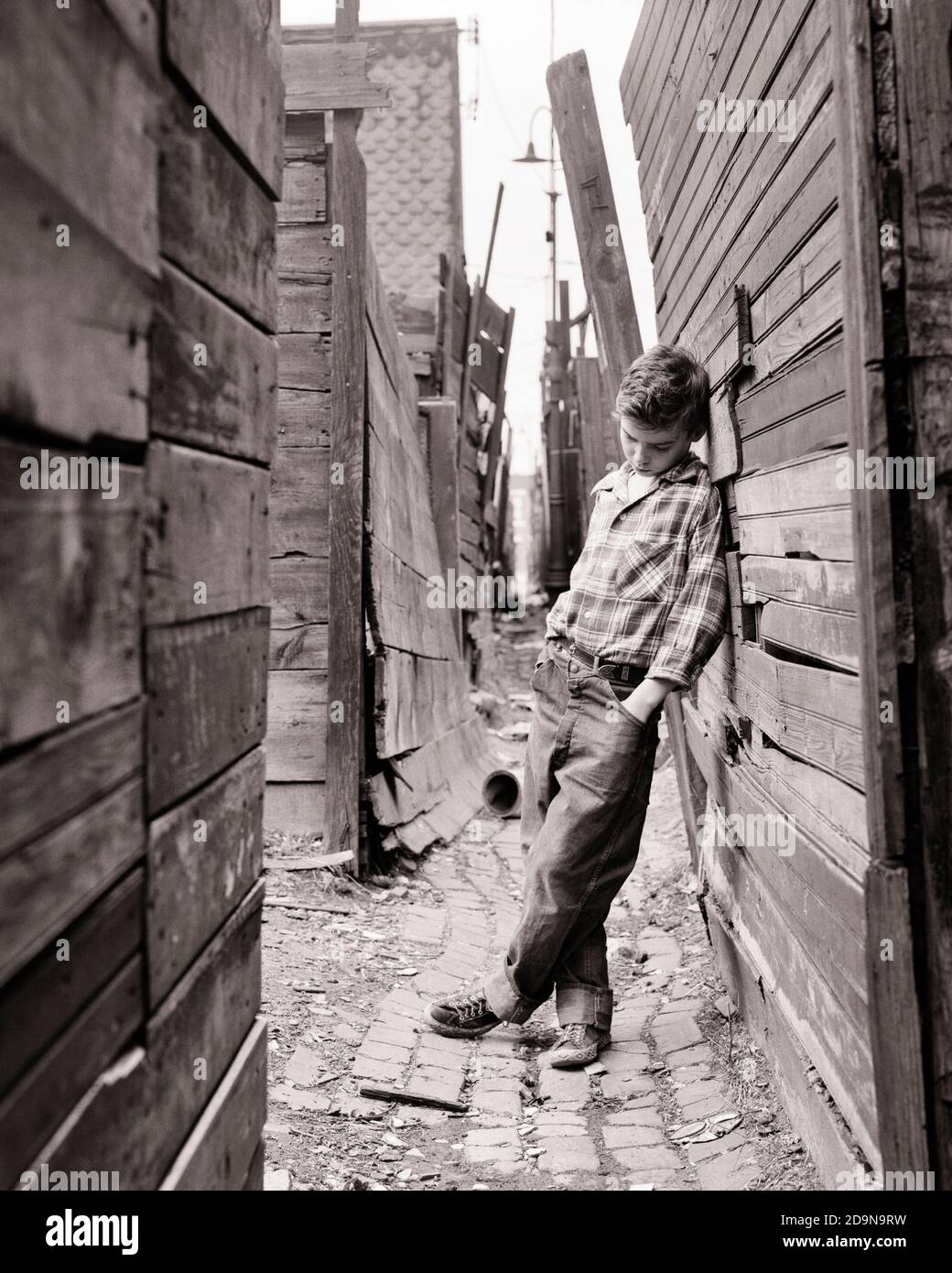 1950s TRUANCY SAD LONELY PRE-TEEN BOY IN POVERTY STRICKEN NEIGHBORHOOD ALLEY LEANING UP AGAINST A WOODEN FENCE PLAYING HOOKY - w1785 HAR001 HARS POOR HOME LIFE COPY SPACE AGAINST FULL-LENGTH MALES RISK DENIM TROUBLED B&W CONCERNED SADNESS DREAMS NEIGHBORHOOD PRIDE UP MOOD GLUM DISADVANTAGED JOYLESS BLUE JEANS DISAFFECTED DISAPPOINTED DISCONNECTED IMPOVERISHED JUVENILES MISERABLE PRE-TEEN PRE-TEEN BOY SLUM STRICKEN BLACK AND WHITE CAUCASIAN ETHNICITY DISTRAUGHT HAR001 NEEDY OLD FASHIONED Stock Photo