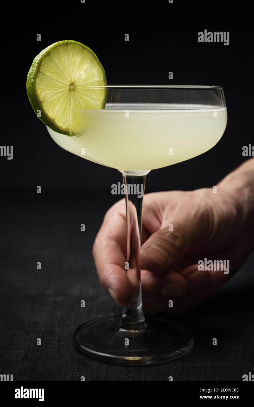 Man's hand grabbing a classic Daiquiri cocktail with a lime wheel from a black table Stock Photo