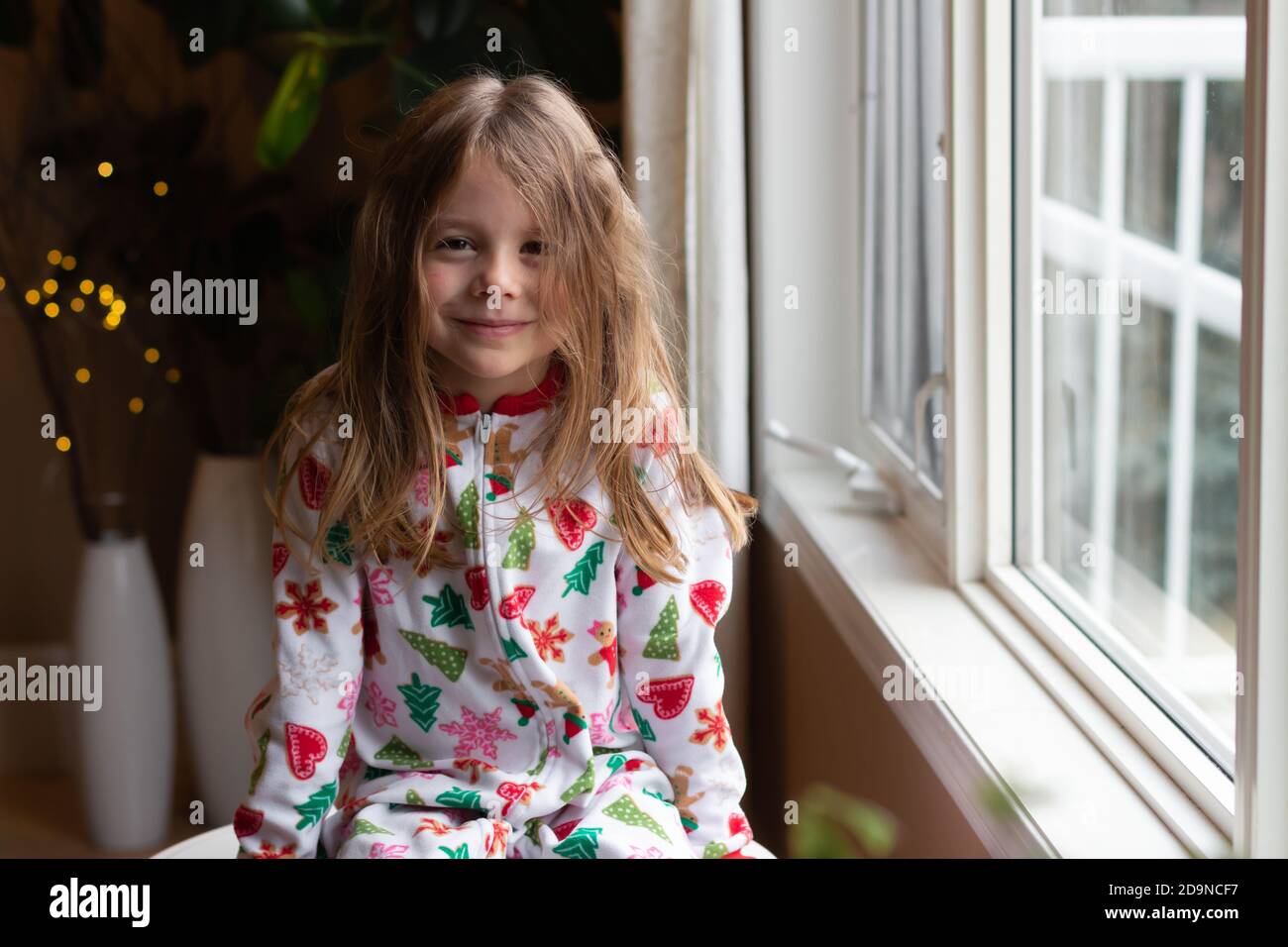 Young girl looking at camera wearing Christmas pyjamas sitting by window Stock Photo