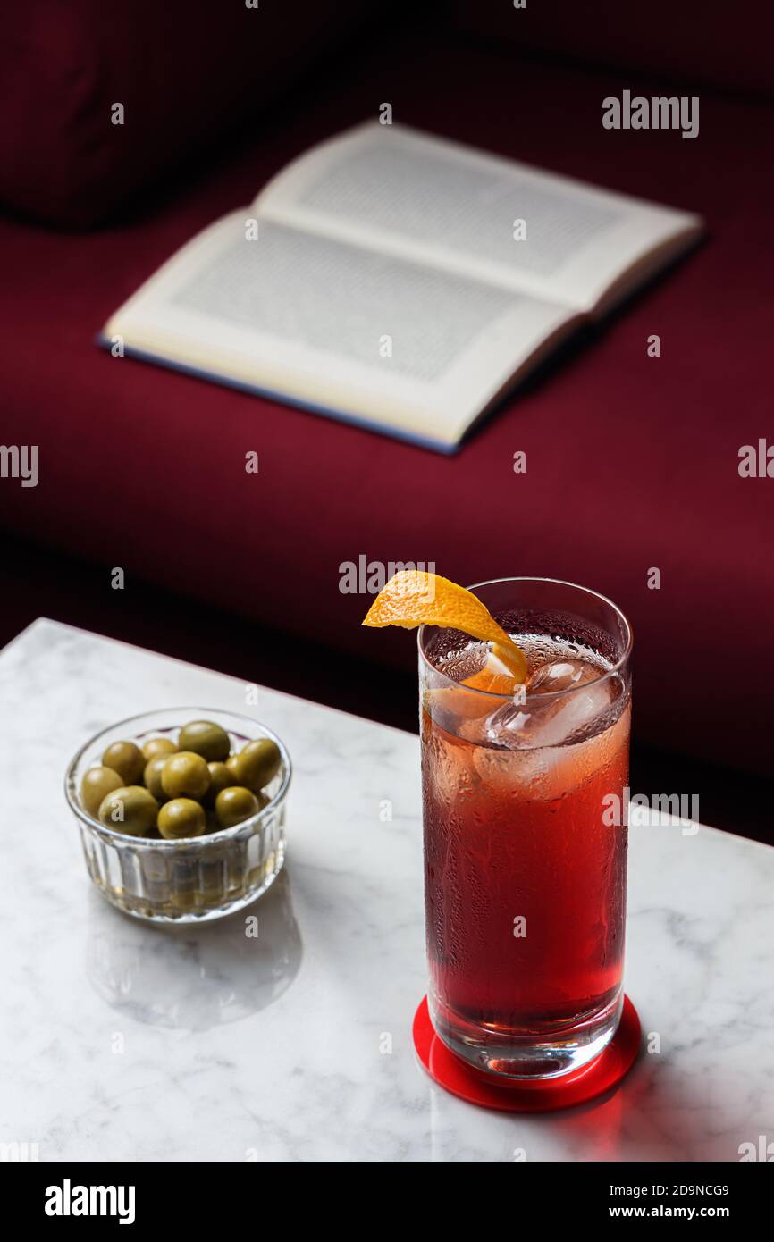 Americano Cocktail and some olives on a marble table. Open book on a red sofa as a background. Relaxing time Stock Photo