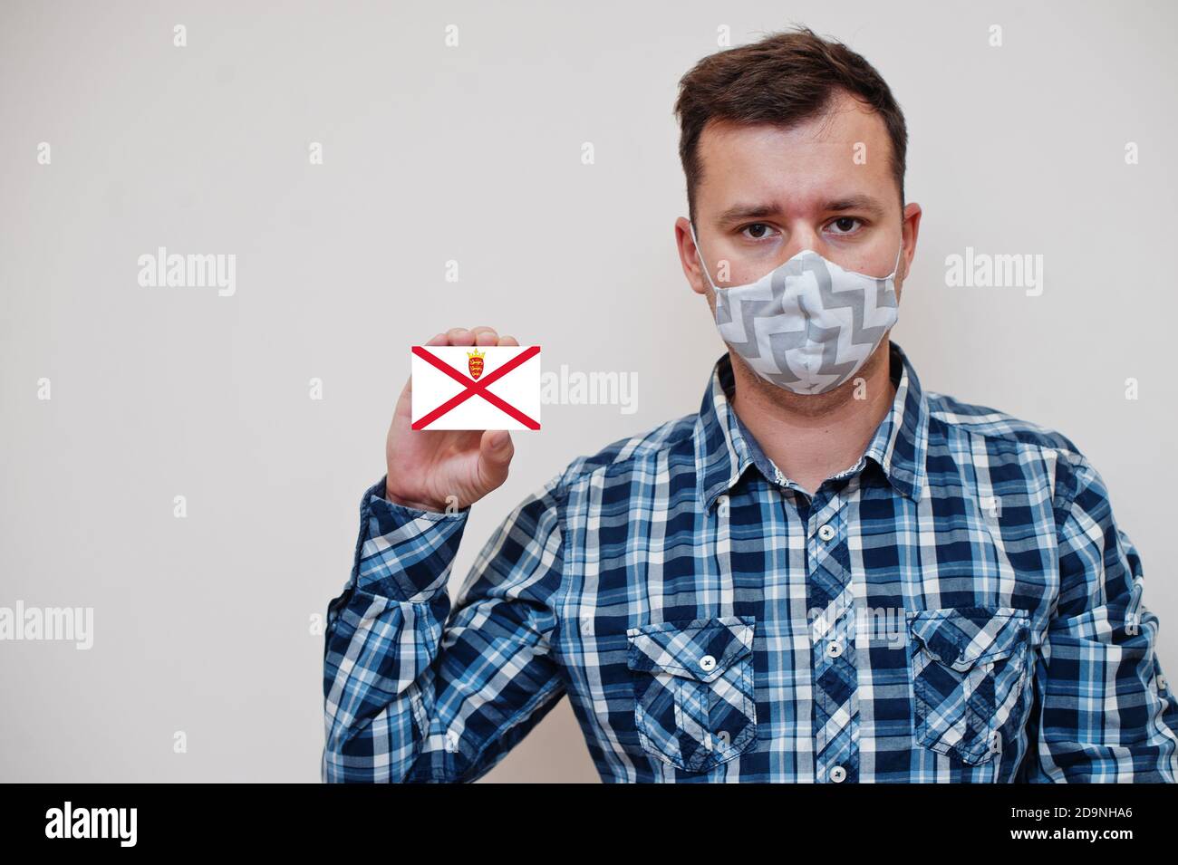 Man in checkered shirt show Bailiwick of Jersey flag card in hand, wear protect mask isolated on white background. Europe countries Coronavirus concep Stock Photo