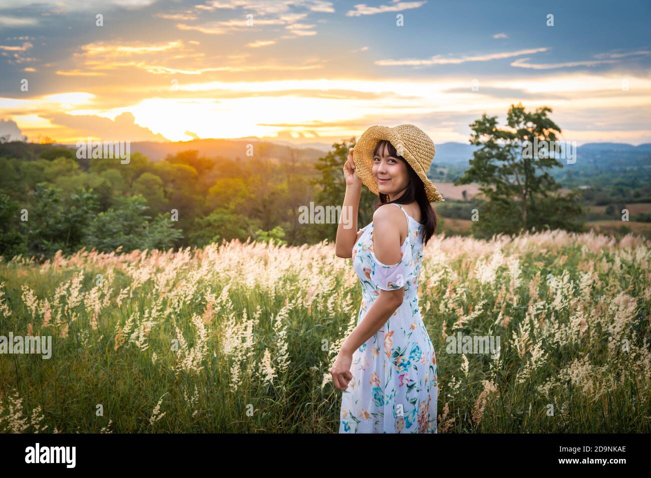 The girl wearing a hat, wearing a white dress, stand in the middle of the grass with beautiful white flowers with a relaxed and happy mood on mountain Stock Photo