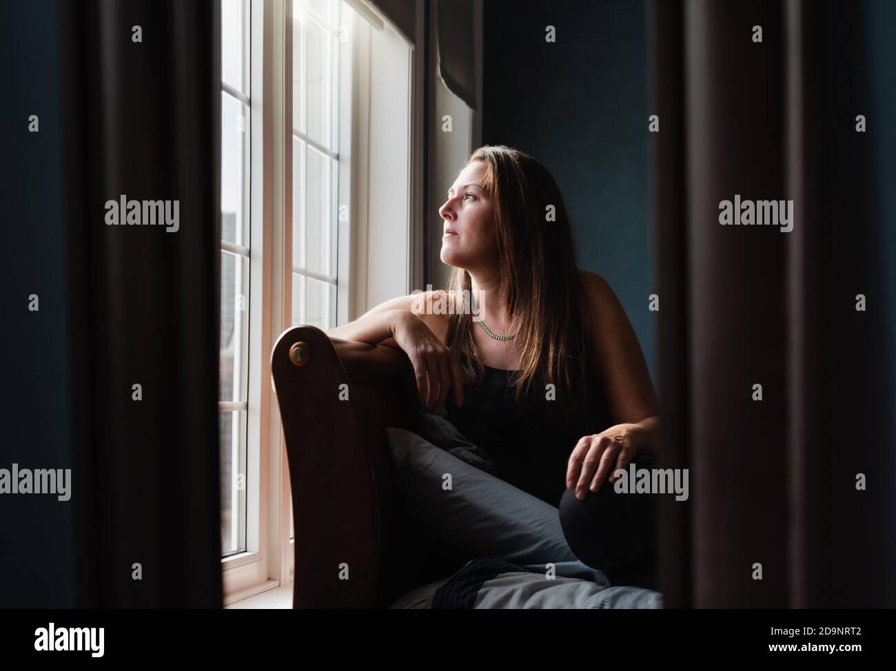 Mirror reflection of pretty woman looking out a window in a dark room ...