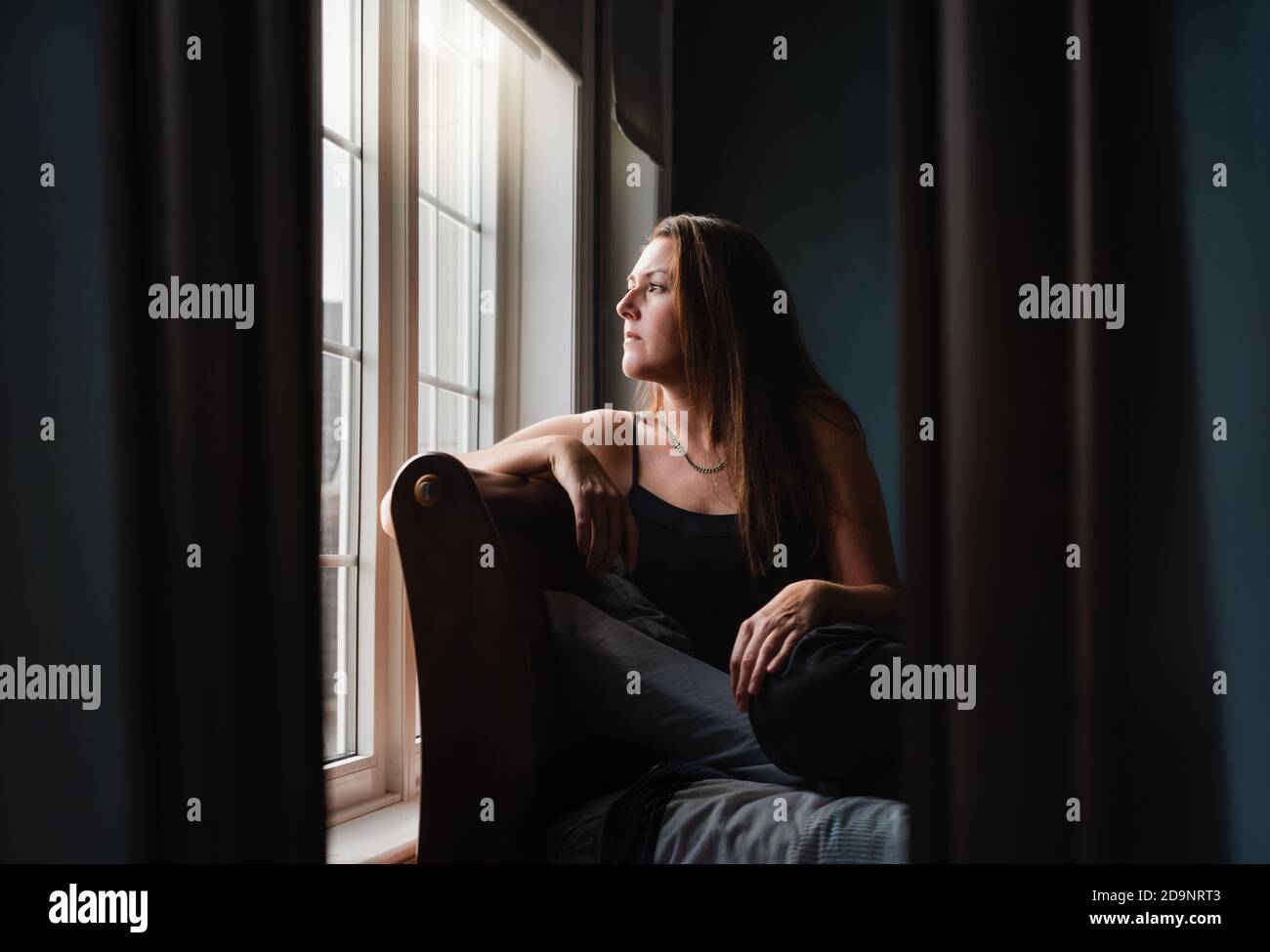 Mirror reflection of pretty woman looking out a window in a dark room ...