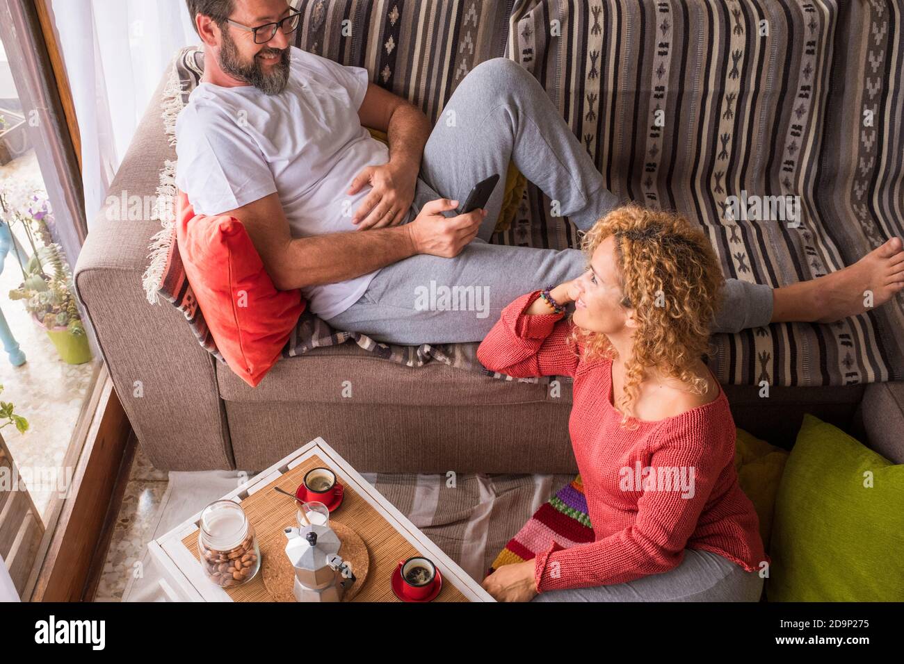 Happy couole enjoy leisure indoor activity at home together with love and relaxed time - man sitting on the couch and wife on the floor having breakfast in the morning - life forever togetherness concept Stock Photo