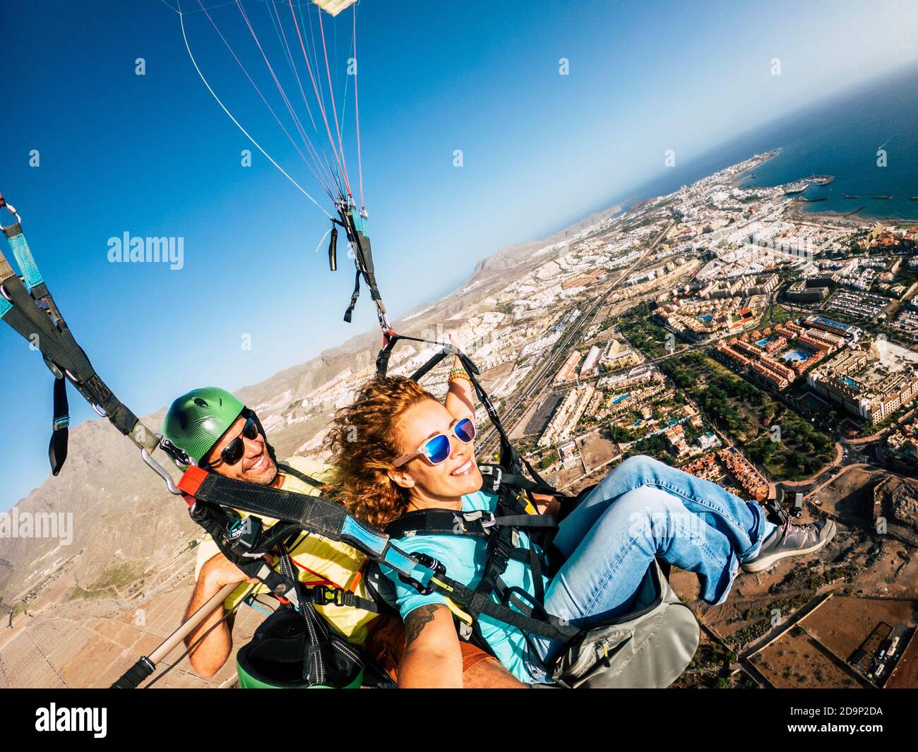 Cheerful happy woman to paraglyde experience with pilot - couple having fun in the air paraglyding over the city with coastline view - tourist and summer holiday vacation active people Stock Photo