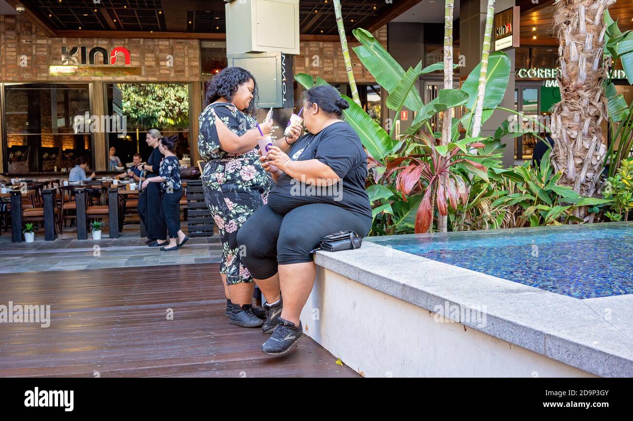 Brisbane, Queensland, Australia - 25th September 2019: Two overweight women drinking a milkshake each as they relax by the pool of a shopping centre Stock Photo