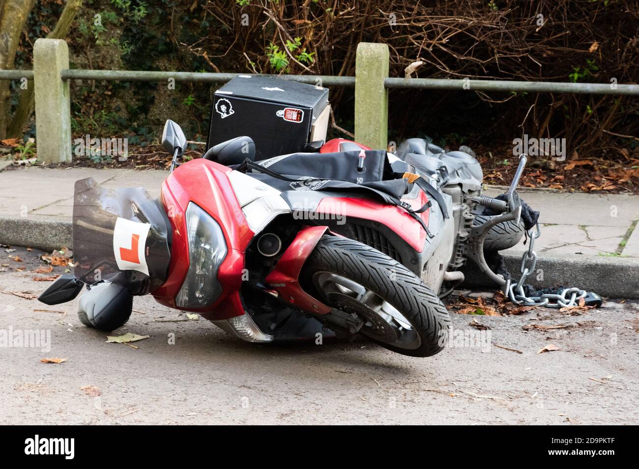 A red moped with ‘L plates’ and security chain that has been tipped onto its side. Stock Photo