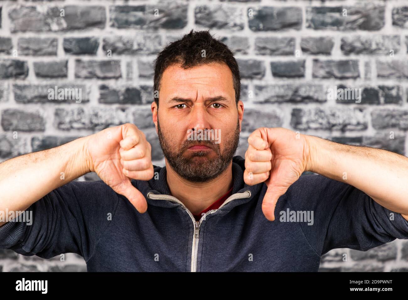 Young caucasian man holding both thumbs down with a frowning face in front of a stone wall Stock Photo
