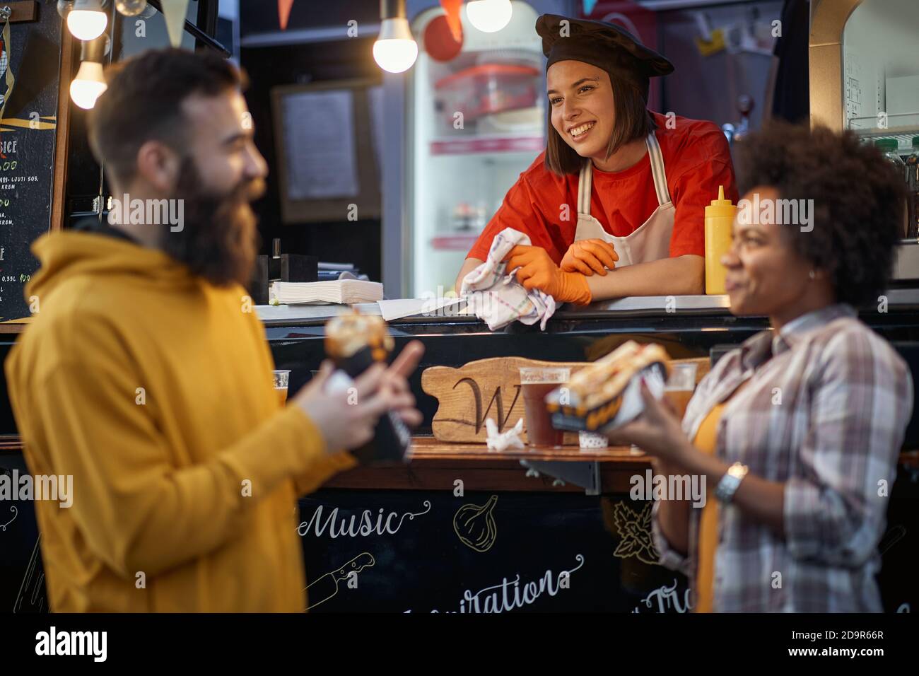 caucasian female employee in fast food service smiling, looking at beardy hipster talking with a male friend Stock Photo