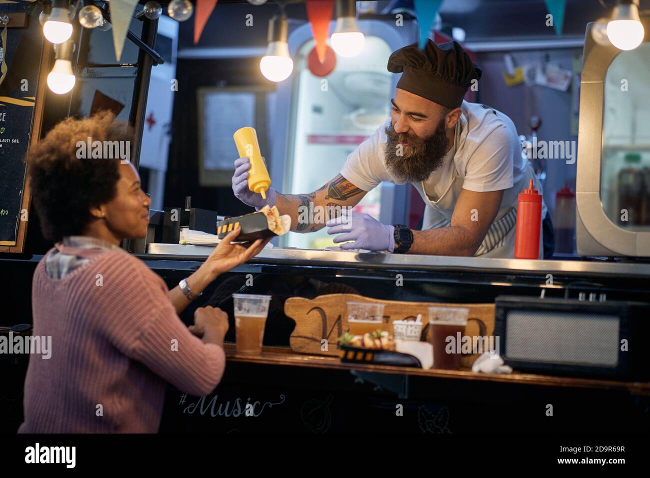young caucasian male employee in fast food service smiling and talking to a afro-american female customer while adding mustard in her sandwich Stock Photo