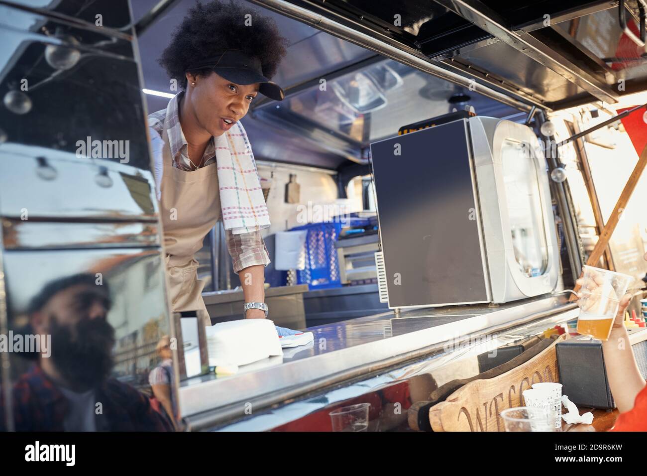 young afro-american female employee in fast food service writing down the order looking at  young caucasian beardy hipster with a cap. Reflection. Stock Photo