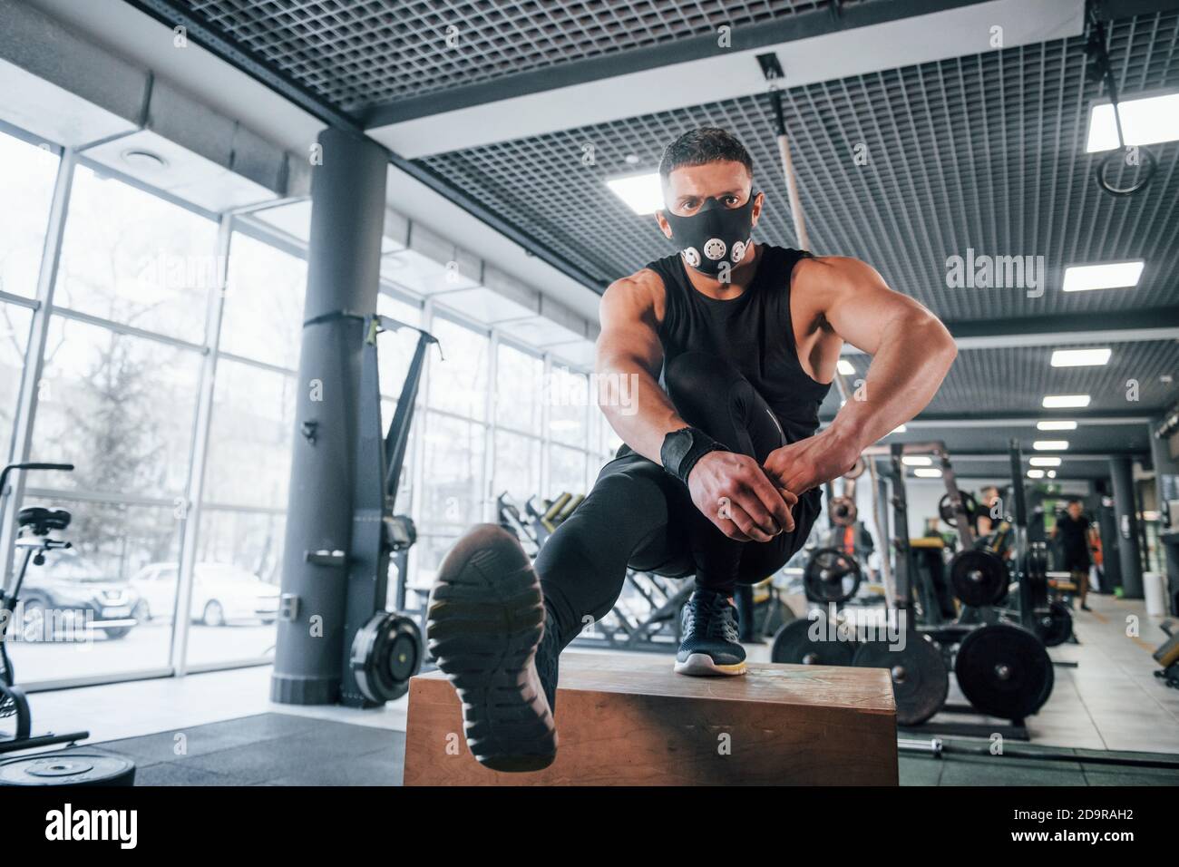 Strong young man in sportive clothes and scary mask doing push ups on one leg in gym Stock Photo