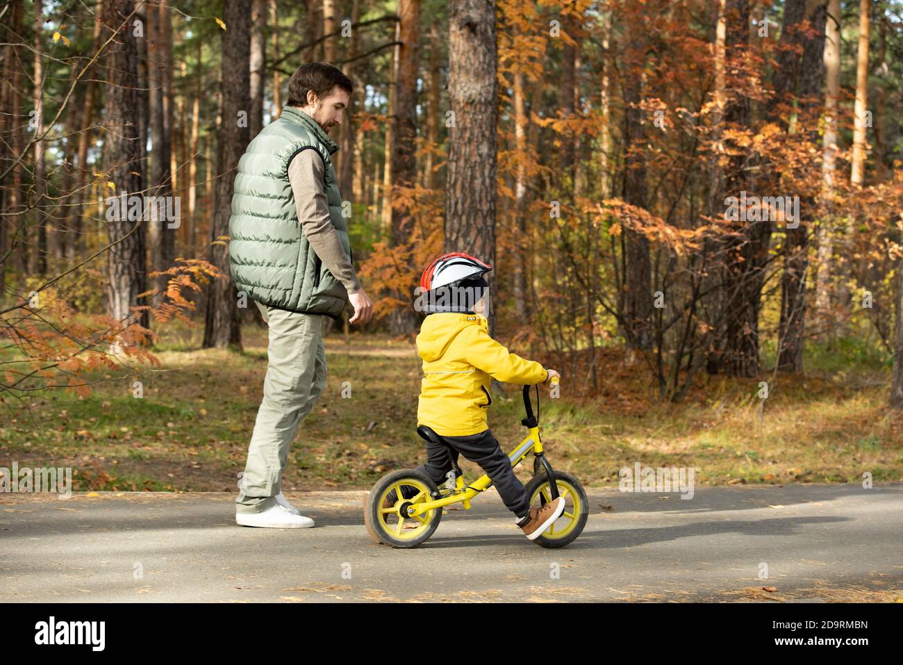 Young man walking by his little son in safety helmet riding balance bicycle Stock Photo