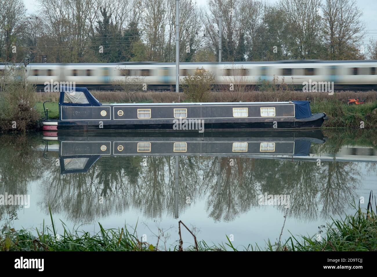 A narrowboat is moored along the River Great Ouse at Buckden in Cambridgeshire, as a commuter train heads along the East Coast mainline. Stock Photo