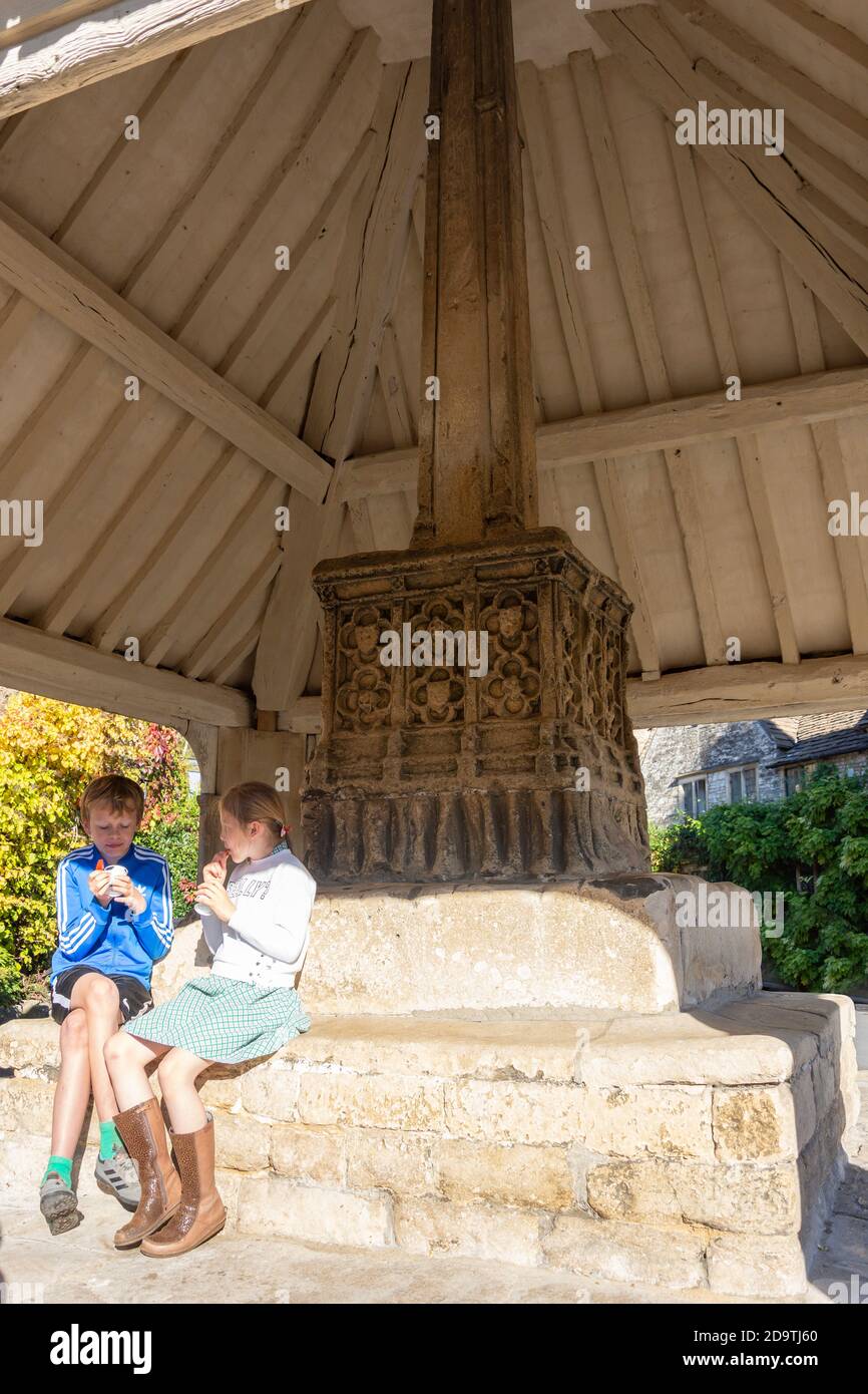 Children sitting by 14th century Market Cross, Market Square, Castle Combe, Wiltshire, England, United Kingdom Stock Photo