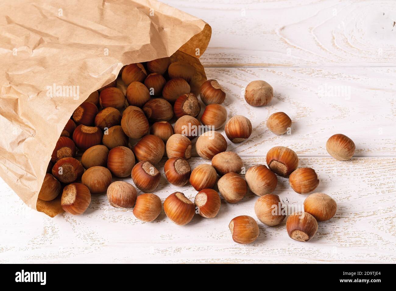 Unpeeled hazelnuts spilled out of a paper bag onto the white wood table. Antioxidant and protein source for ketogenic and raw food diets. Stock Photo