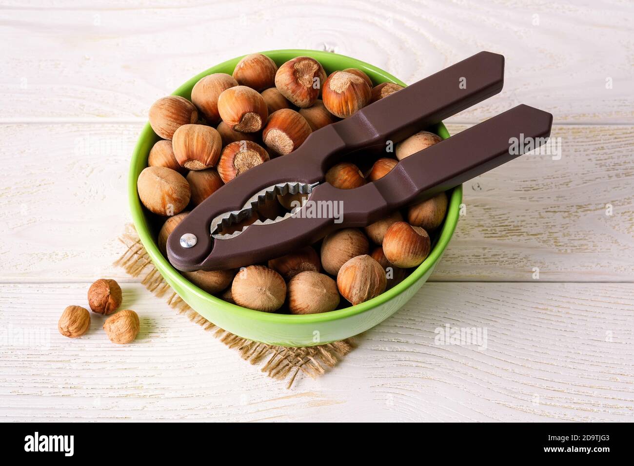Brown nut cracker on a green bowl full of unpeeled hazelnuts over white wood table. Antioxidant and protein source for ketogenic and raw food diets. Stock Photo